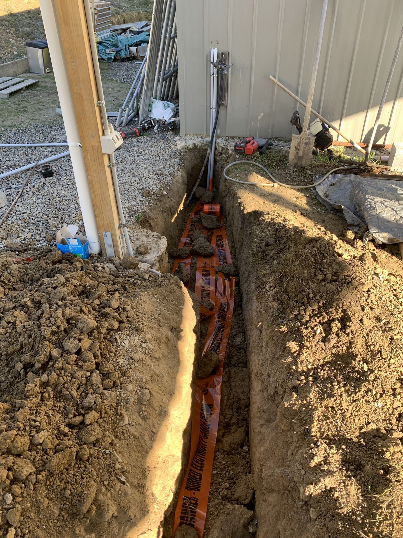 Open trench with orange buried electricity cable marker tape running to a steel shed